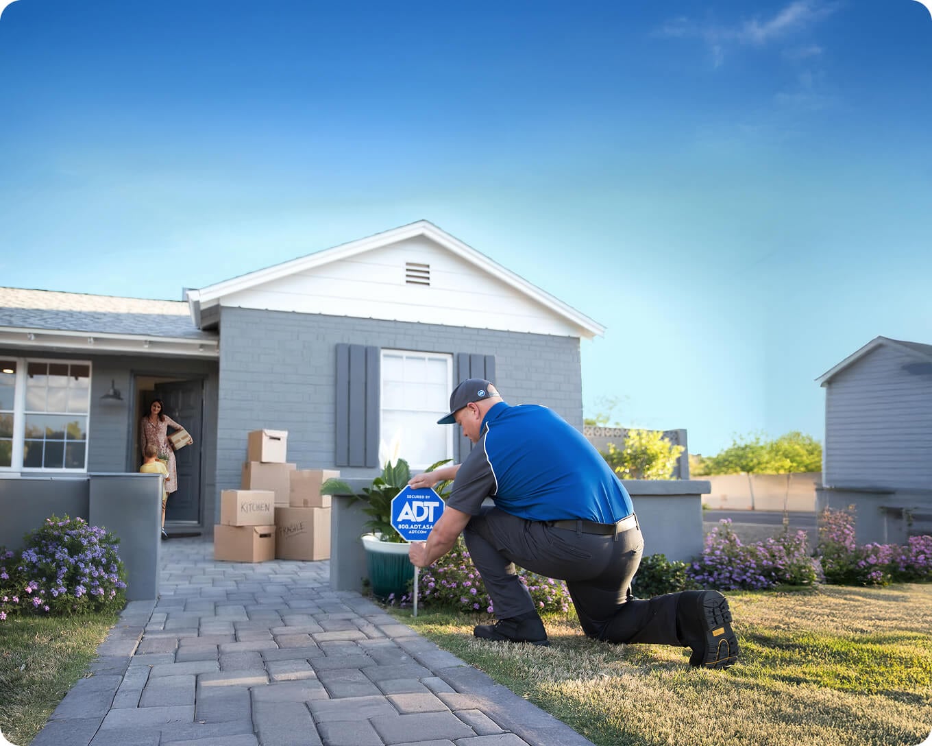 Técnico profesional de instalación de ADT instalando el cartel de jardín de ADT fuera de la casa de ladrillo mientras el nuevo propietario está en el umbral con cajas de mudanza.